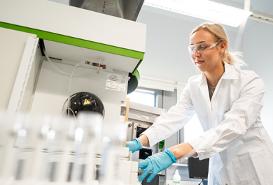 Nuori nainen työskentelee laboratoriossa yllään valkoinen labratakki, suojalasit ja kädessä siniset hanskat. Kva otettu alaviistosta ja etualalla näkyy koeputkia