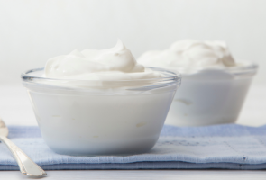 Pöydällä on lusikka ja kaksi lasikippoa, joissa on kukkuralliset annokset valkoista jogurttia.