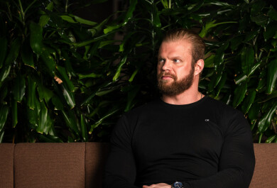 Lauri Vasankari istuu ruskealla sohvalla, takana tummanvihreätapettinen seinä. Henkilöllä yllään musta neule ja farkut, jalat ojentuneena eteen nilkat ristikkäin, kädet sylissä ja katsoo vasemmalle