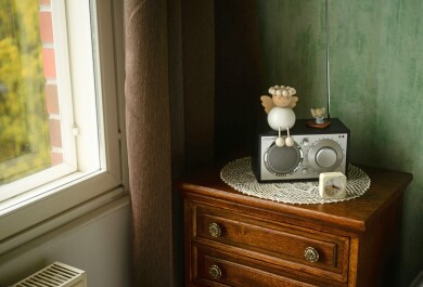 Huoneen nurkka, jossa lipasto. Lipaston päällä pitsiliina, radio, enkelikoristeita ja herätyskello. Toisella seinustalla ikkuna.