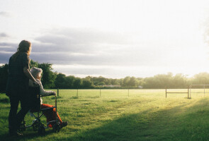 Kuvituskuvassa nuorempi henkilö työntää vanhempaa henkilöä pyörätuolissa kauniissa maisemassa