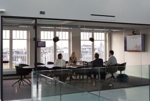 Neljä ihmistä pitää kokousta pöydän ääressä tilassa, jonka seinät ovat lasia.