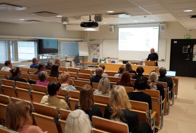 Ihmiset kuuntelevat tilan edessä puhuvaa naista.