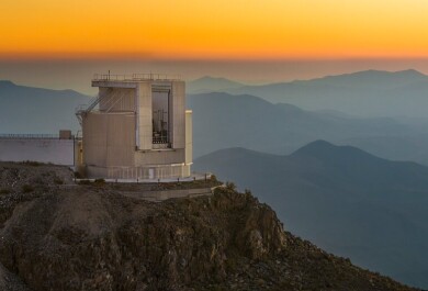 Valokuva observatioriosta vuoren rinteellä, taustalla vuoria ja oranssia taivasta.