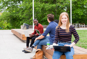 Kolme nuorta ihmistä istuu Yliopistonmäen penkeillä aurinkoisena päivänä.