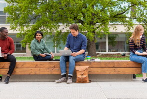 Neljä henkilöä istuu puisella penkillä ulkona, taustalla rakennus ja puu.