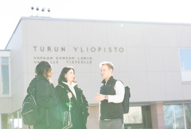 Kolme henkilöä juttelemassa iloisesti Turun yliopiston päärakennuksen edessä. Taustalla yliopiston päärakennuksen seinä, jossa inskriptio "vapaan kansan lahja vapaalle tieteelle".